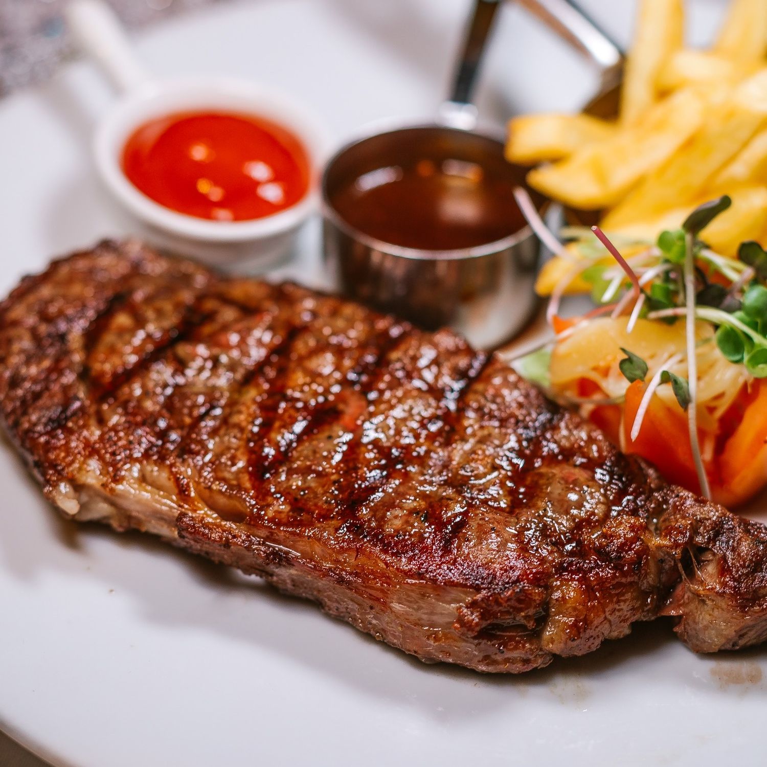 A grilled New York strip steak served with French fries, cherry tomatoes and two dipping sauces on a white plate.