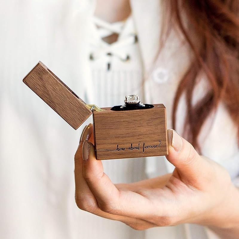 Small wooden proposal ring box with flip-top lid being held in hands against white background.