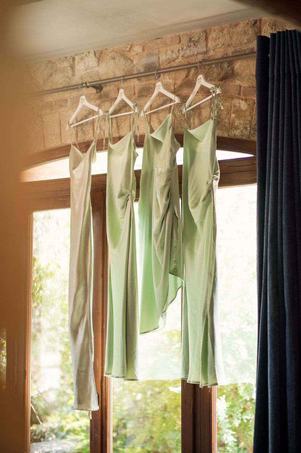Mint green and neutral bridesmaid dresses hanging on white hangers against a stone wall.
