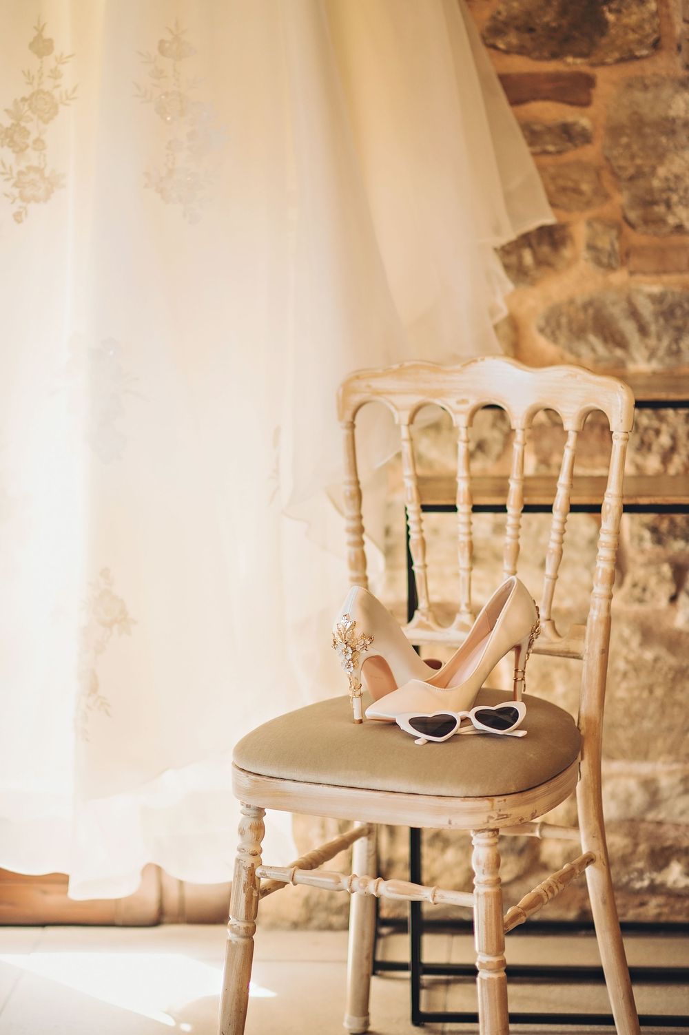 Wedding accessories including sunglasses and heels arranged on a vintage chair against stone walls.