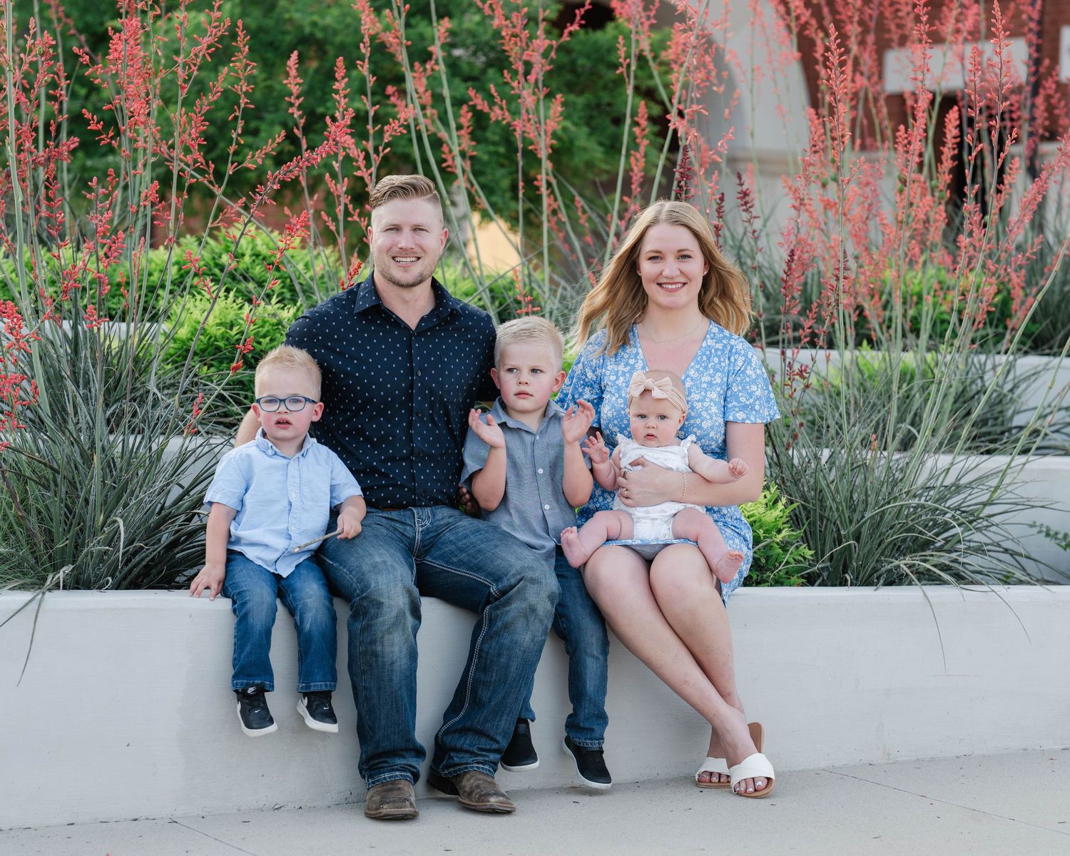 A family sits together outdoors on a concrete bench with ornamental grasses and pink flowers behind them.