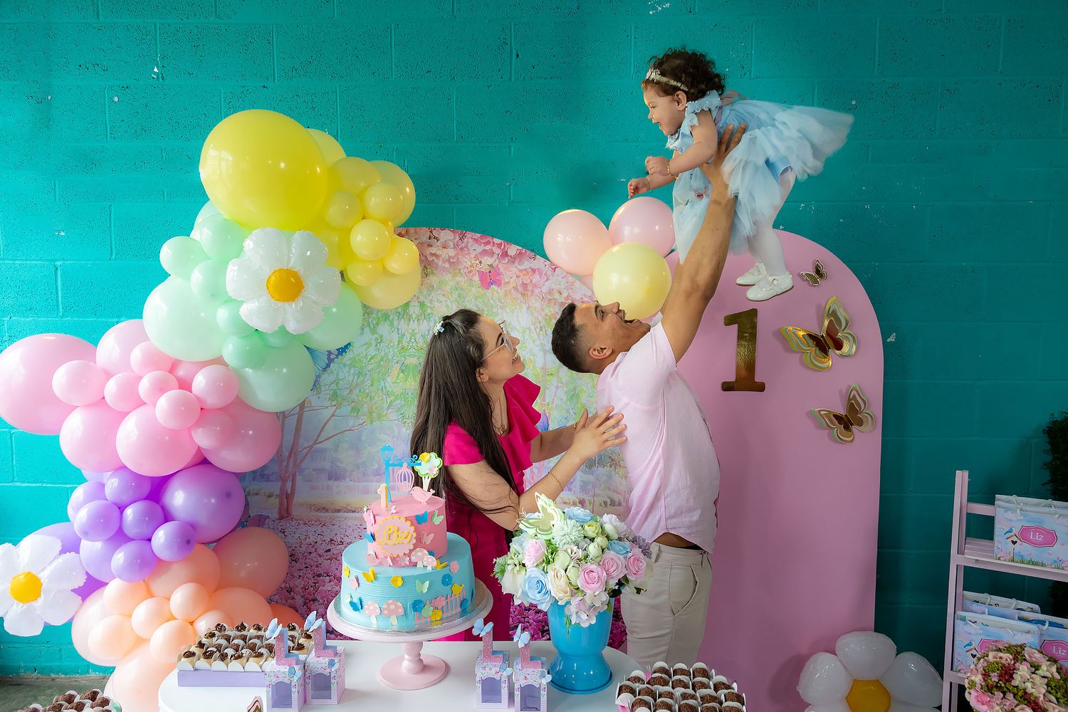 People setting up colorful balloon decorations and cake display for a milestone celebration against turquoise wall.
