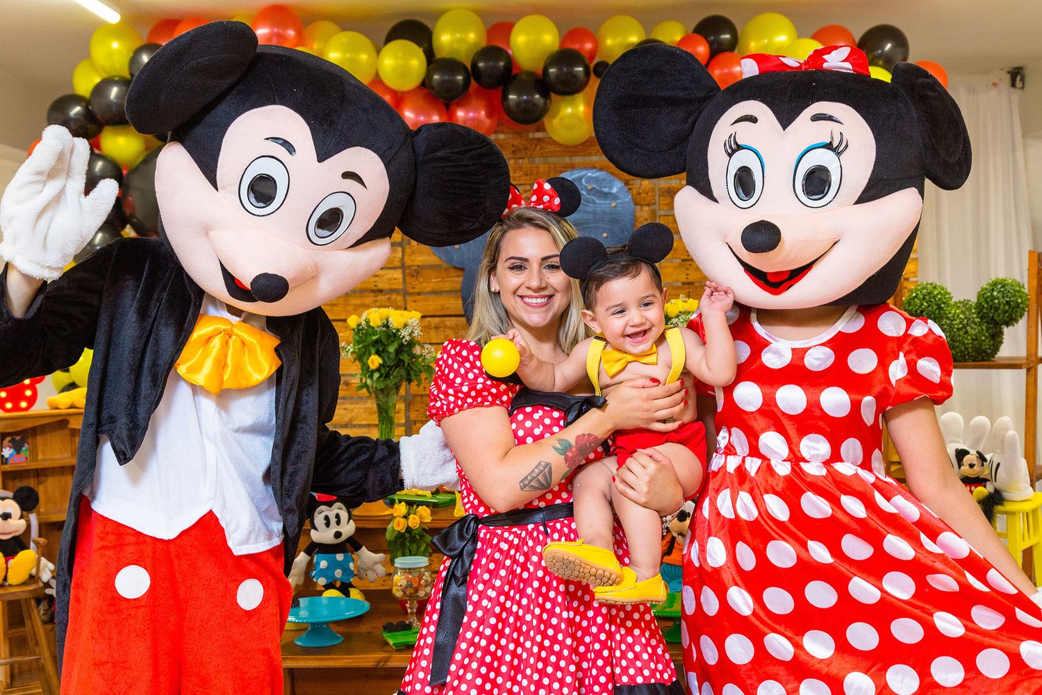 A festive party scene with Mickey and Minnie Mouse characters in costumes posing with guests against colorful balloon decorations.