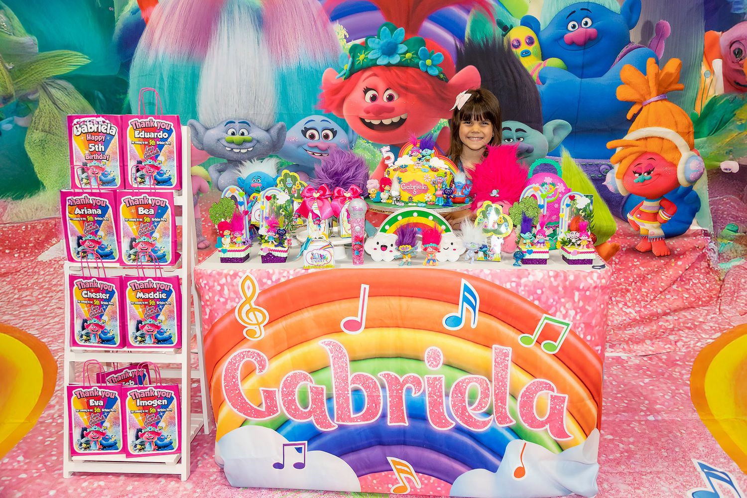 A colorful Trolls-themed birthday party setup with decorations and party favors arranged on a pink table backdrop.