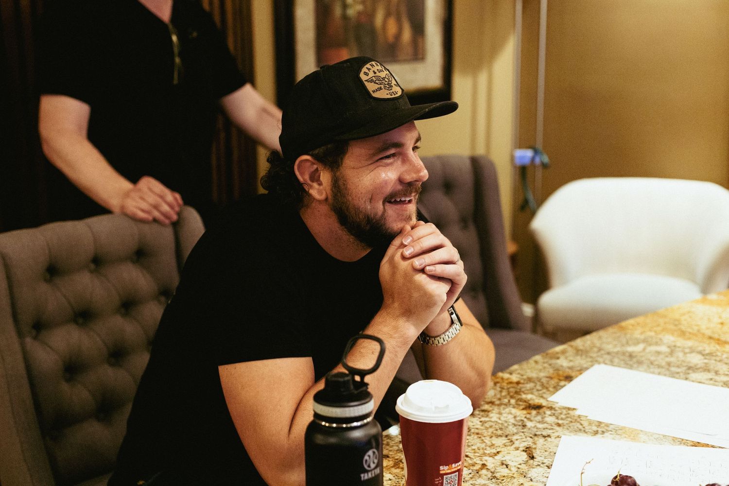 Music artist Matt Jordan wearing a baseball cap smiles while sitting at a table with a drink in a recording studio.