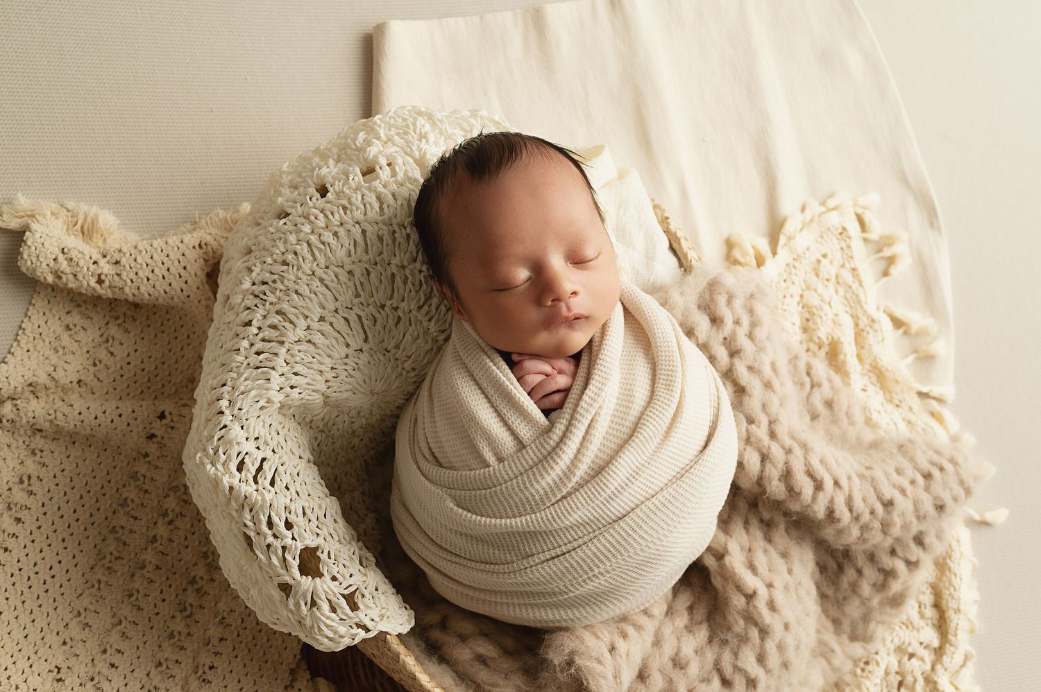 Sleeping newborn wrapped in cream fabric on textured knit blanket.