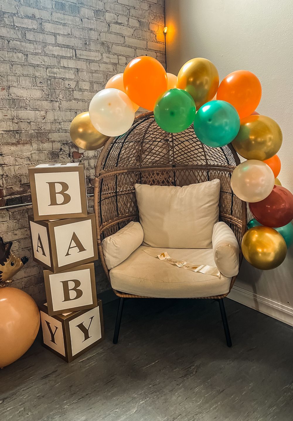 Baby shower or nursery setup with BABY letter blocks, a cozy egg chair, and colorful balloon garland against a brick wall.