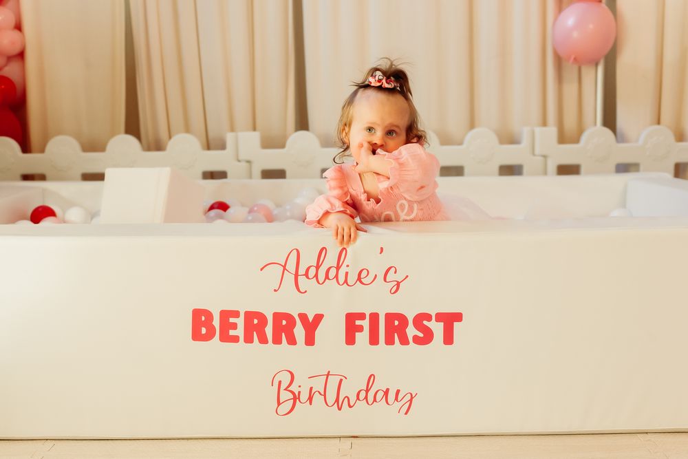 A white crib decorated with pink text reading 'Berry First Birthday' and pink balloons for a themed celebration.
