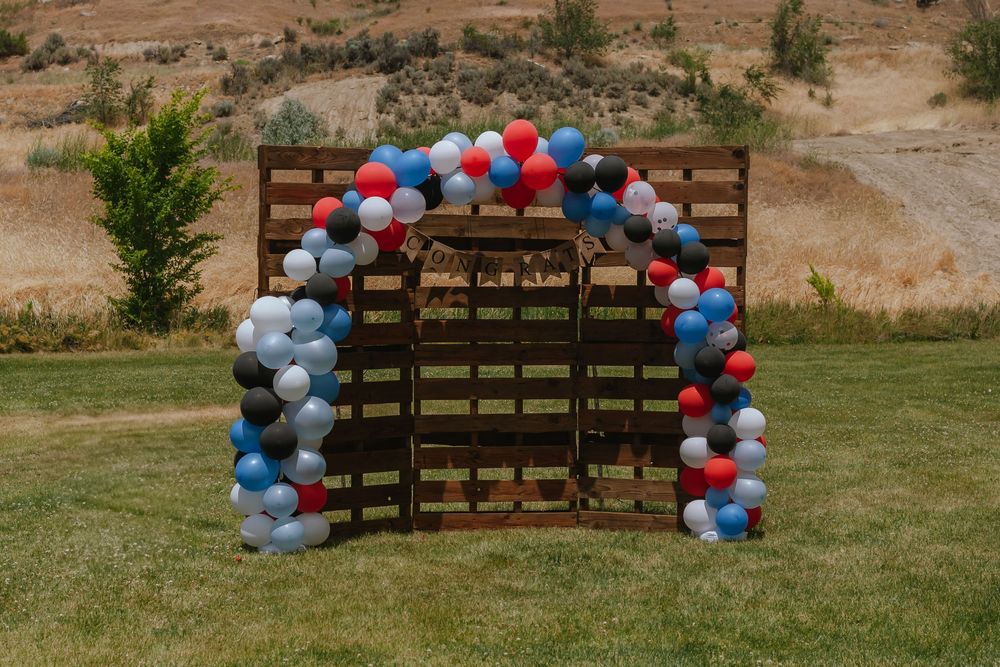 A wooden backdrop arch decorated with red, white, and blue balloon garland stands on grass against a hillside backdrop.
