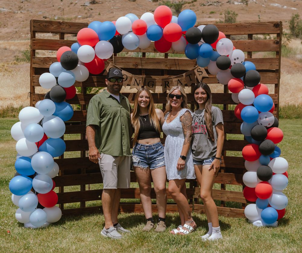 A festive red, white and blue balloon arch frames a group posing in front of a wooden pallet backdrop.