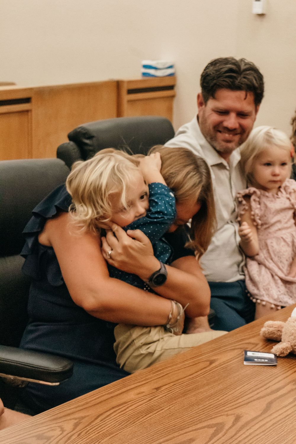 A candid family moment shows parents sharing laughter with their young children around a wooden table.