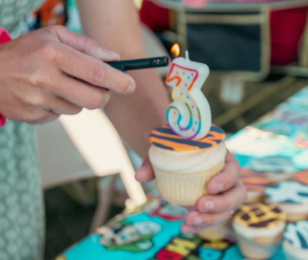 Someone holding a birthday cupcake with a number 3 candle being lit against a blue tablecloth background.