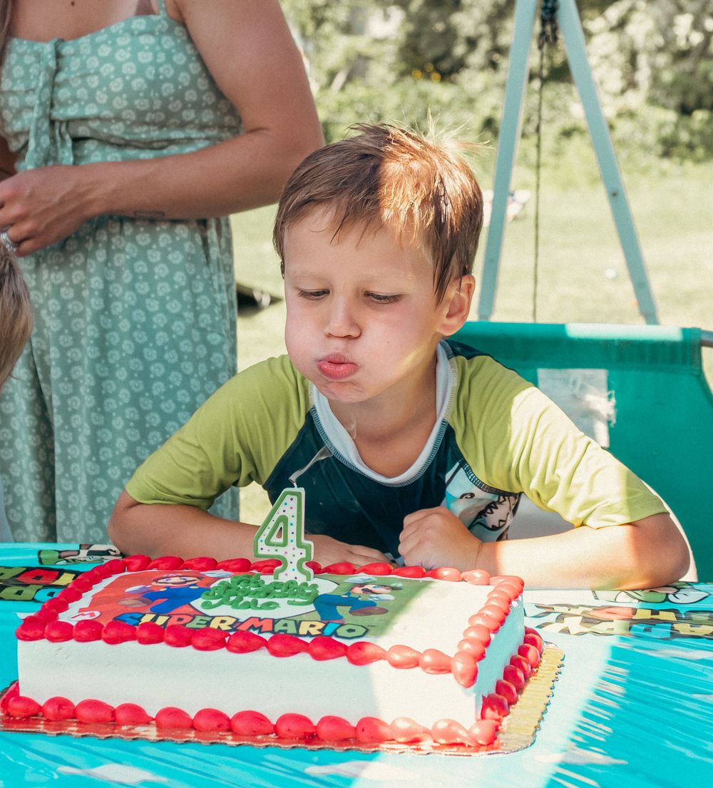 A young child wearing a yellow shirt blows out a number 4 candle on a birthday cake at an outdoor party.