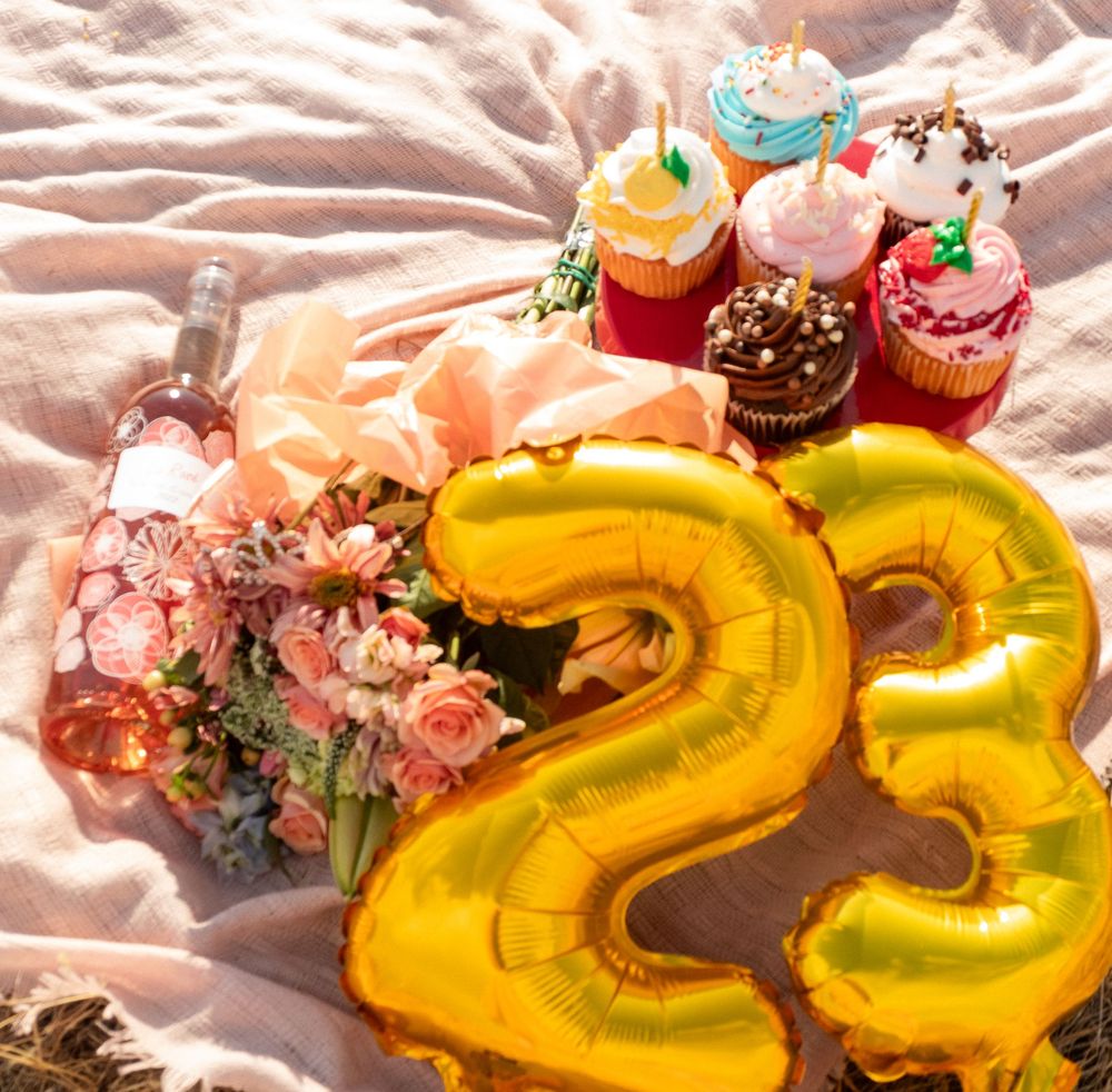 Golden number 23 balloon displayed with colorful cupcakes and pink flowers on pink fabric background.