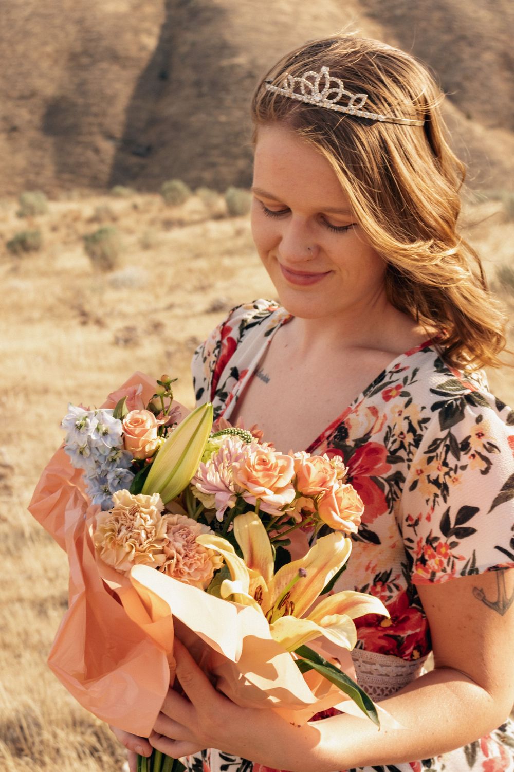 Someone wearing a tiara and floral dress smiles while holding a beautiful bouquet of peach roses and lilies against desert hills.