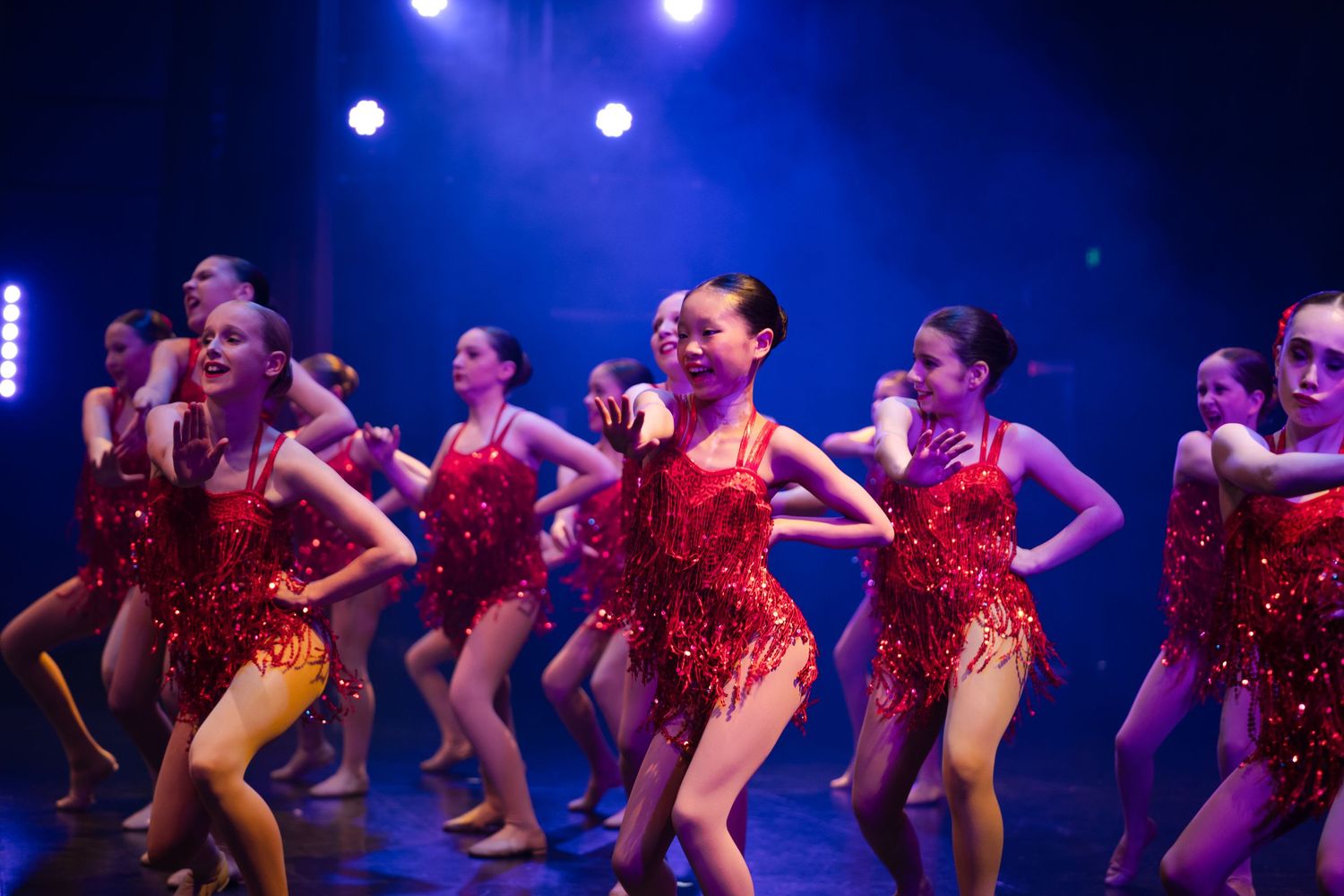 Dancers in sparkling red costumes perform on stage under blue theatrical lighting.