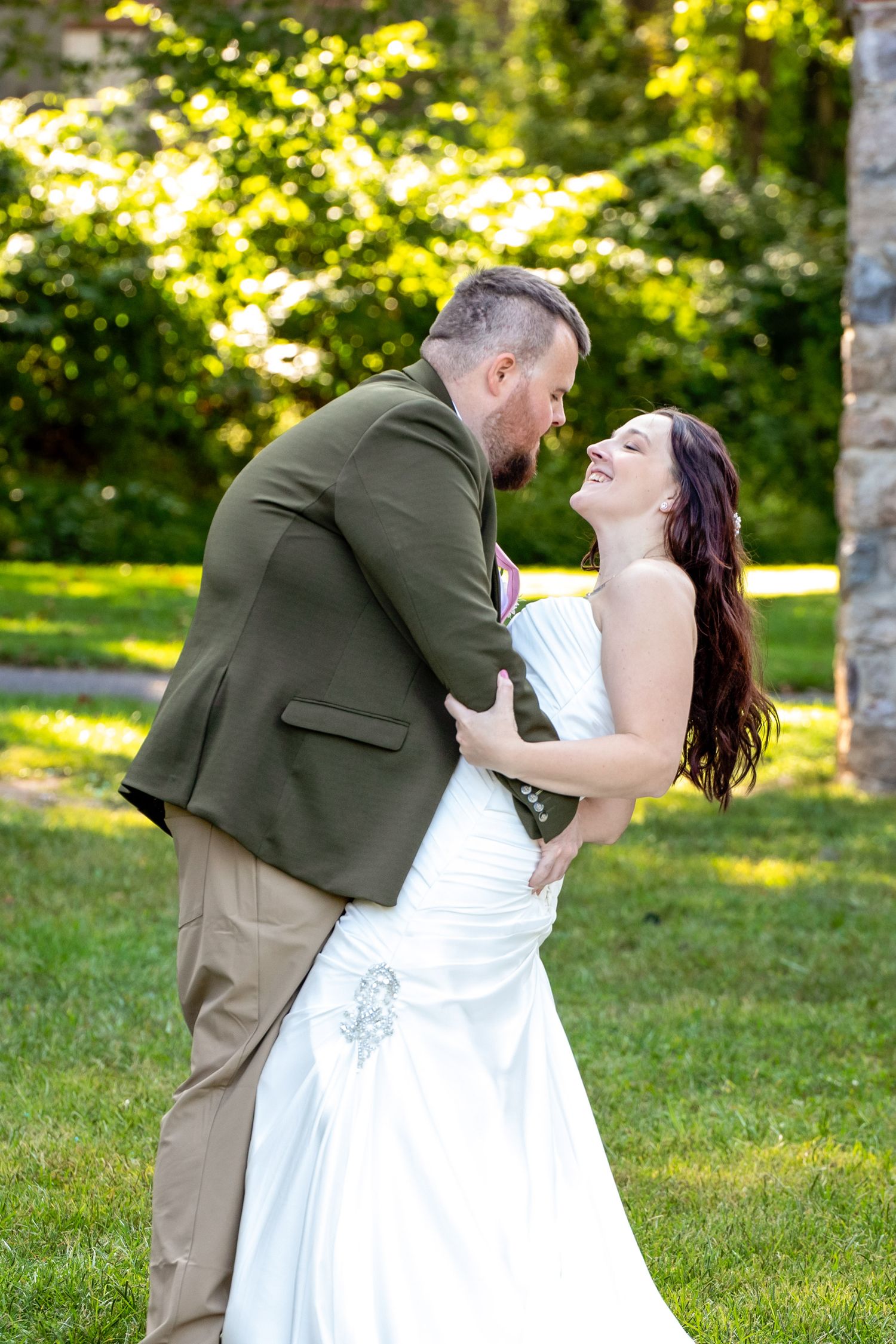 A romantic wedding couple dances together outdoors in a sunlit garden with green trees in the background.