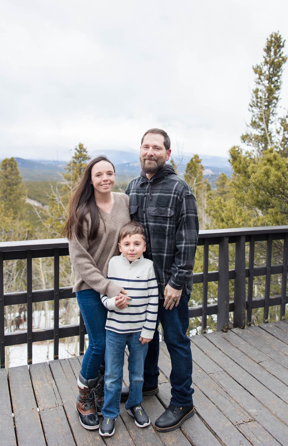 A family poses together on a wooden deck overlooking a mountain landscape with evergreen trees in the background.