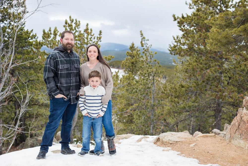 Three people bundled up in winter clothing stand together on a snowy overlook with mountain pine trees in the background.