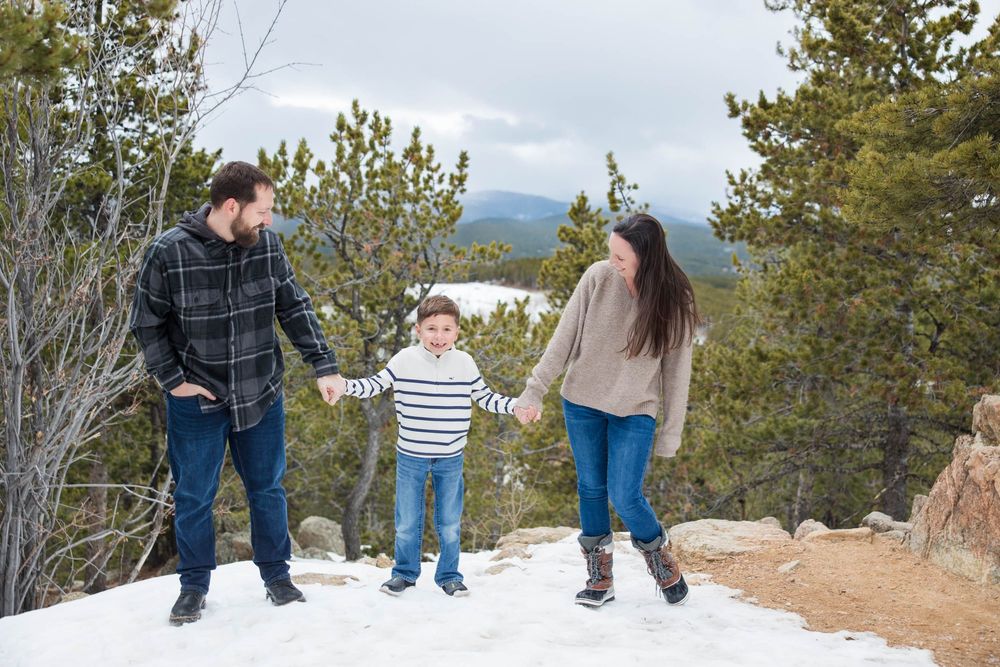 A family stands together on a snow-covered mountain path with evergreen trees in the background.