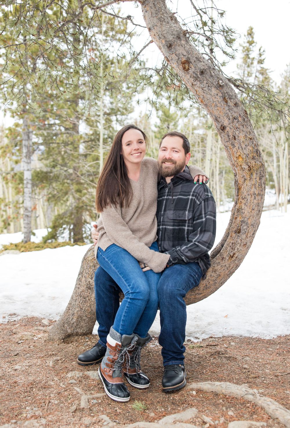 A couple sits together on a curved tree trunk with snow and winter forest scenery behind them.
