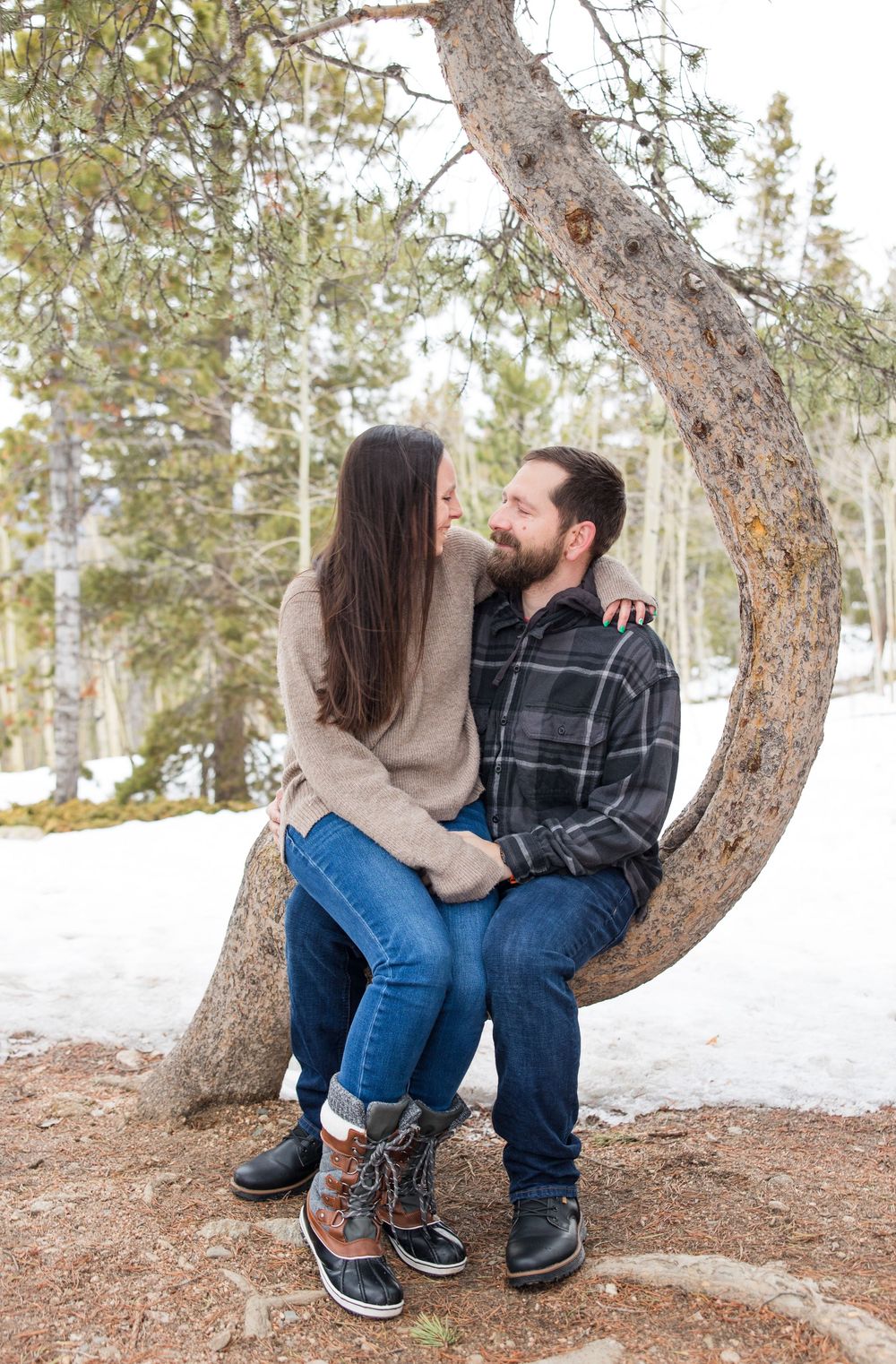 A couple sits together on a curved tree trunk in a snowy mountain setting wearing winter clothing.