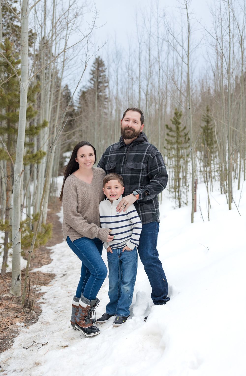A family poses together on a snowy winter path surrounded by bare aspen trees and evergreens in a forest setting.