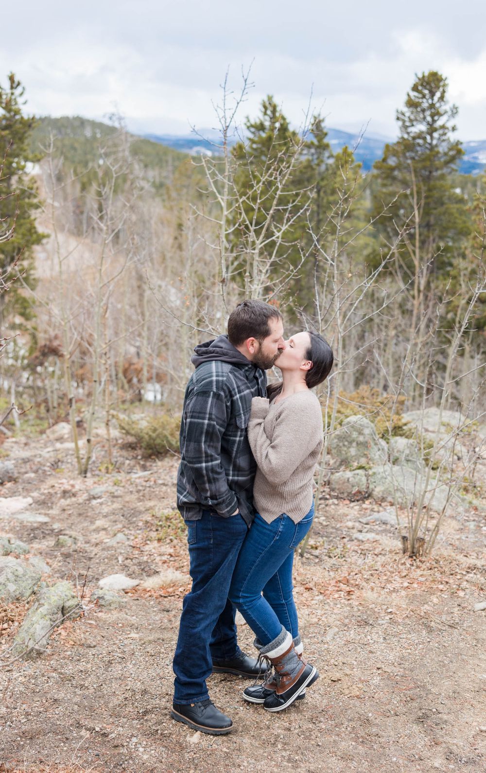 A couple shares a romantic moment while standing on a mountain trail surrounded by winter trees and distant peaks.