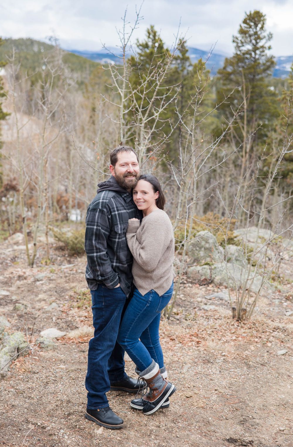Two people embrace while standing on a mountain trail surrounded by bare trees and evergreens in winter.