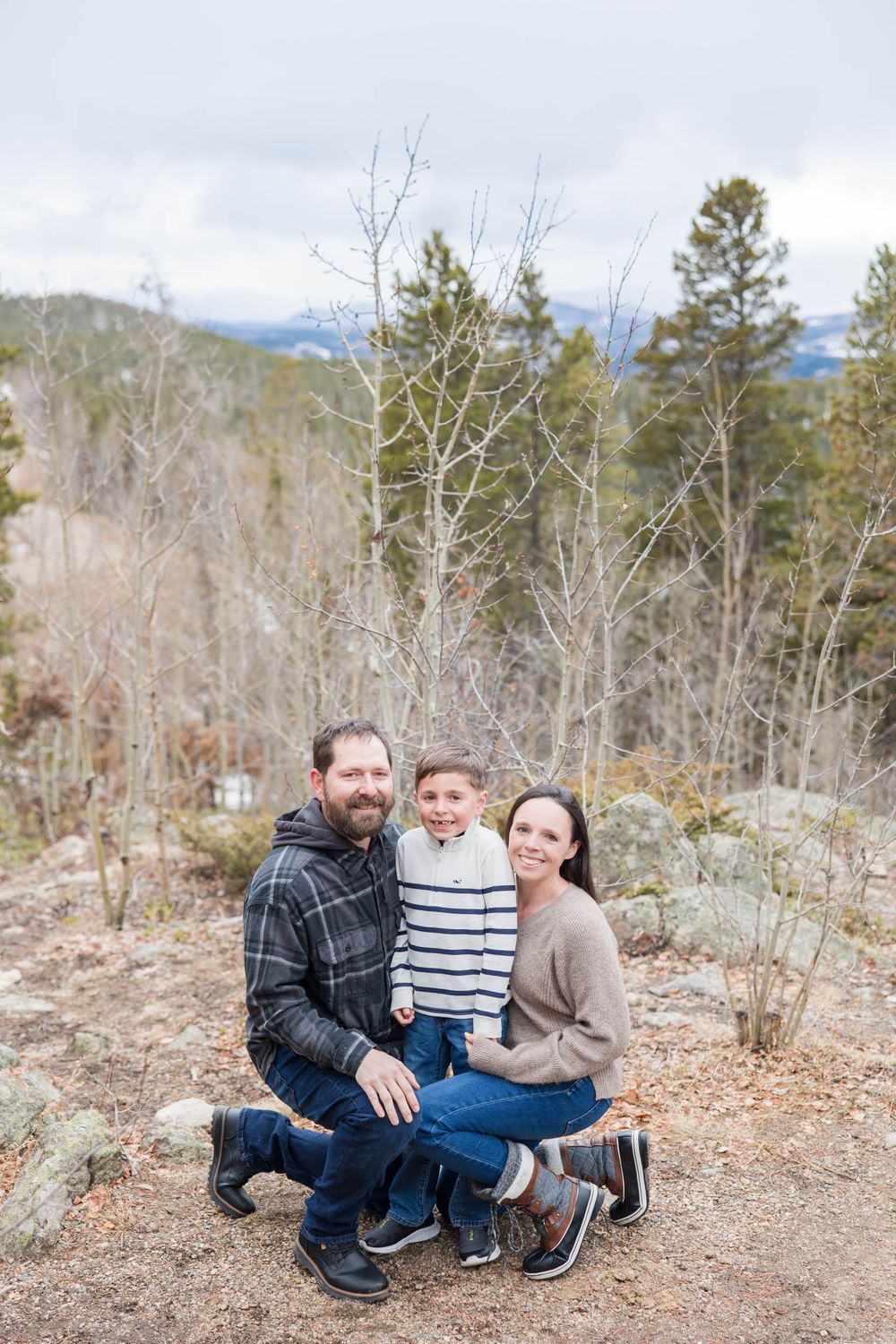 Family poses for outdoor portrait in mountain landscape with bare trees and evergreens in late fall scenery.