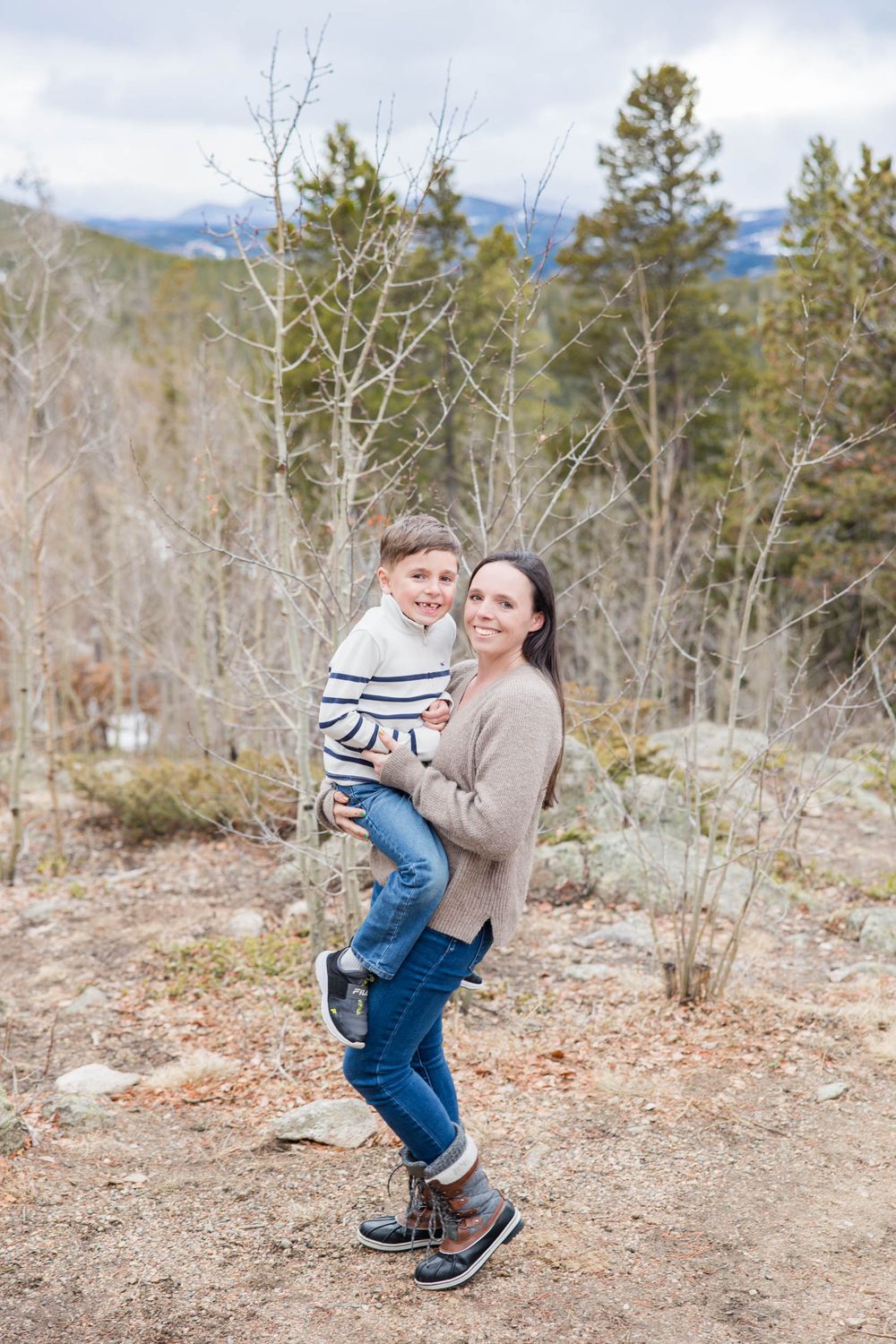 A parent and child pose together on a mountain trail surrounded by bare winter trees and evergreens.