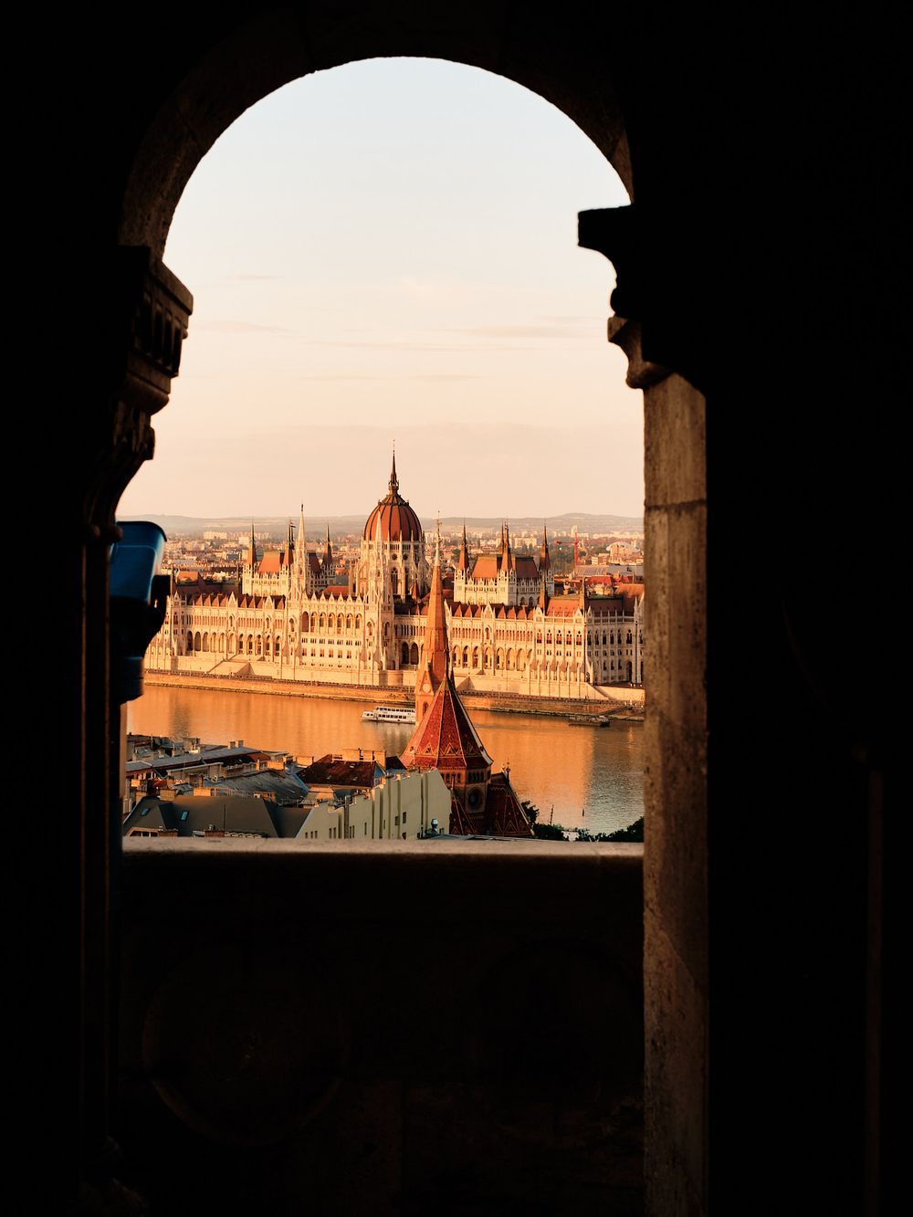 View of the Hungarian Parliament Building through an arch at sunset along the Danube River in Budapest.