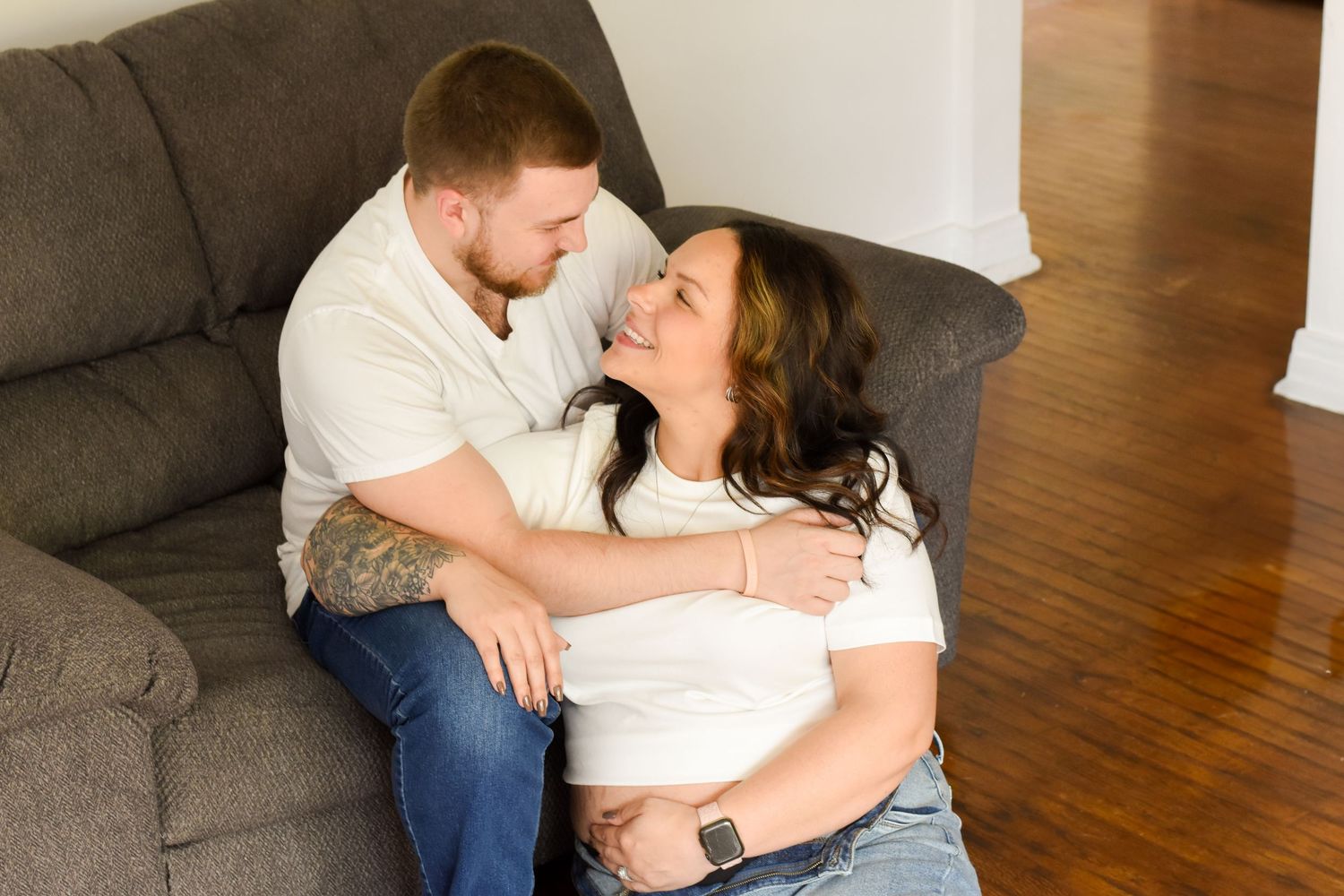 A couple shares an intimate moment while relaxing together on a grey couch in a room with hardwood floors.