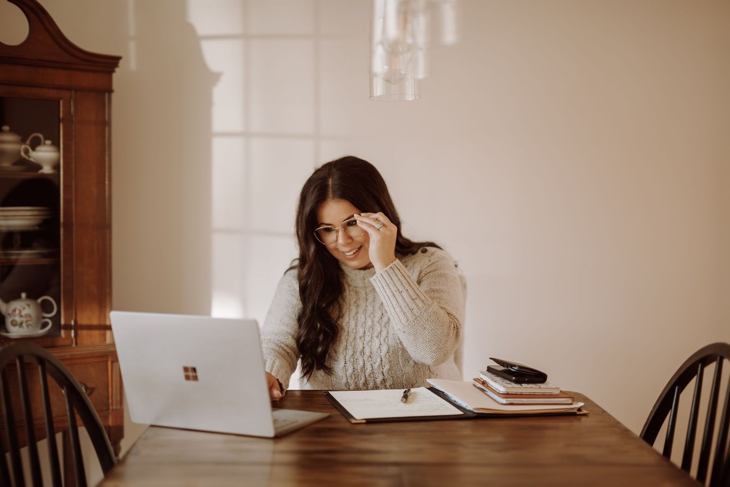 A person in a cream sweater works on a Microsoft Surface laptop at a wooden table with natural lighting streaming in.