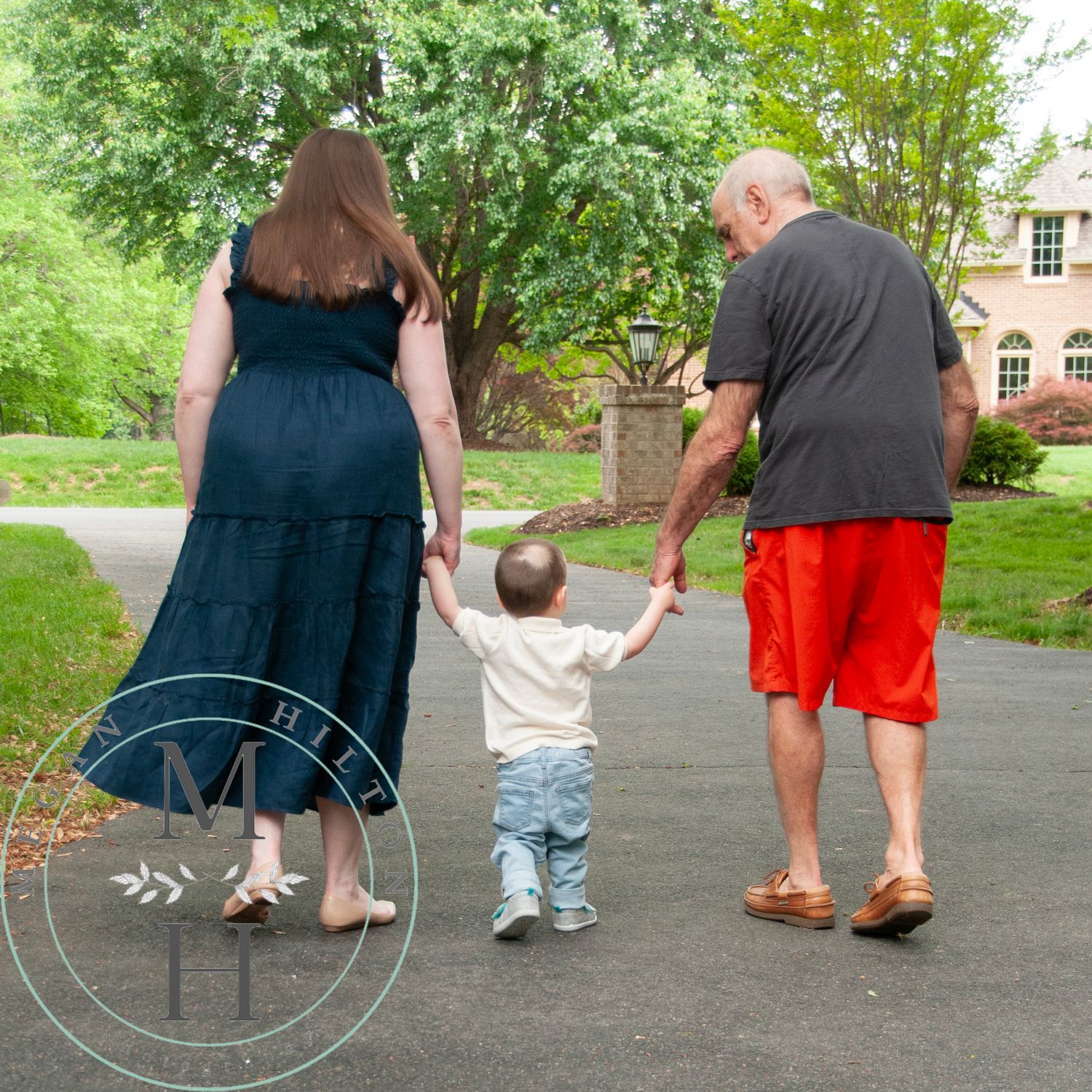 A small child holds hands with two adults while walking together on a paved path in a green garden setting.