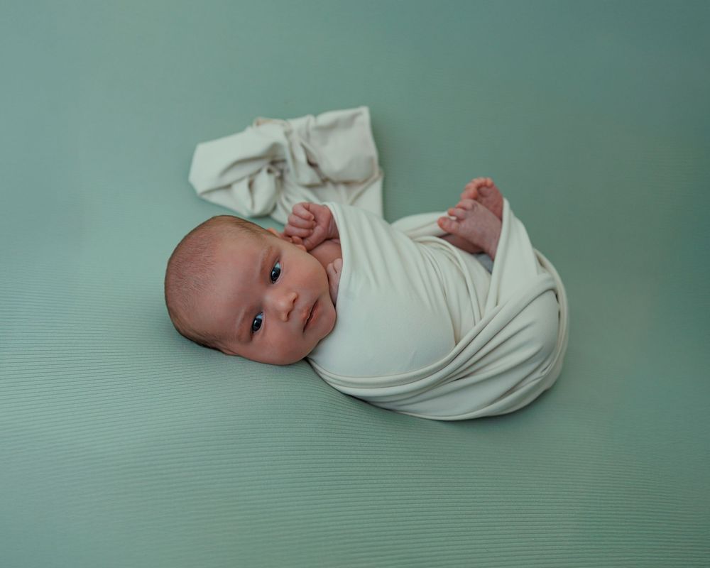 A newborn baby swaddled in white fabric lying on a mint green background.