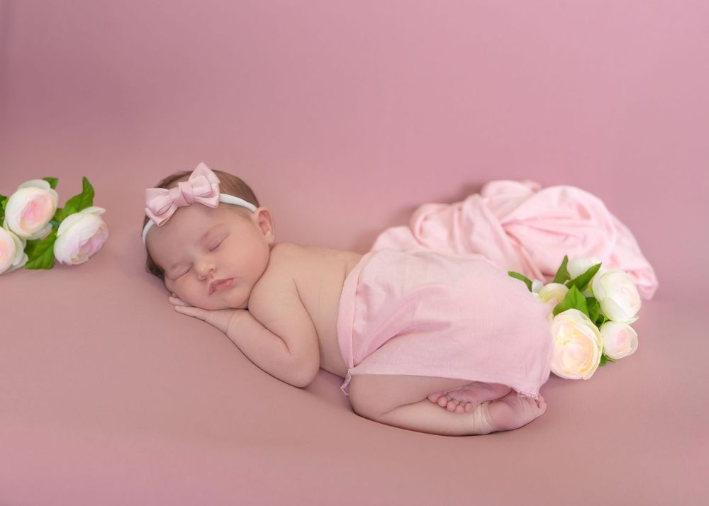 Sleeping newborn in pink wrap lies peacefully on pink backdrop surrounded by delicate white and pink roses.