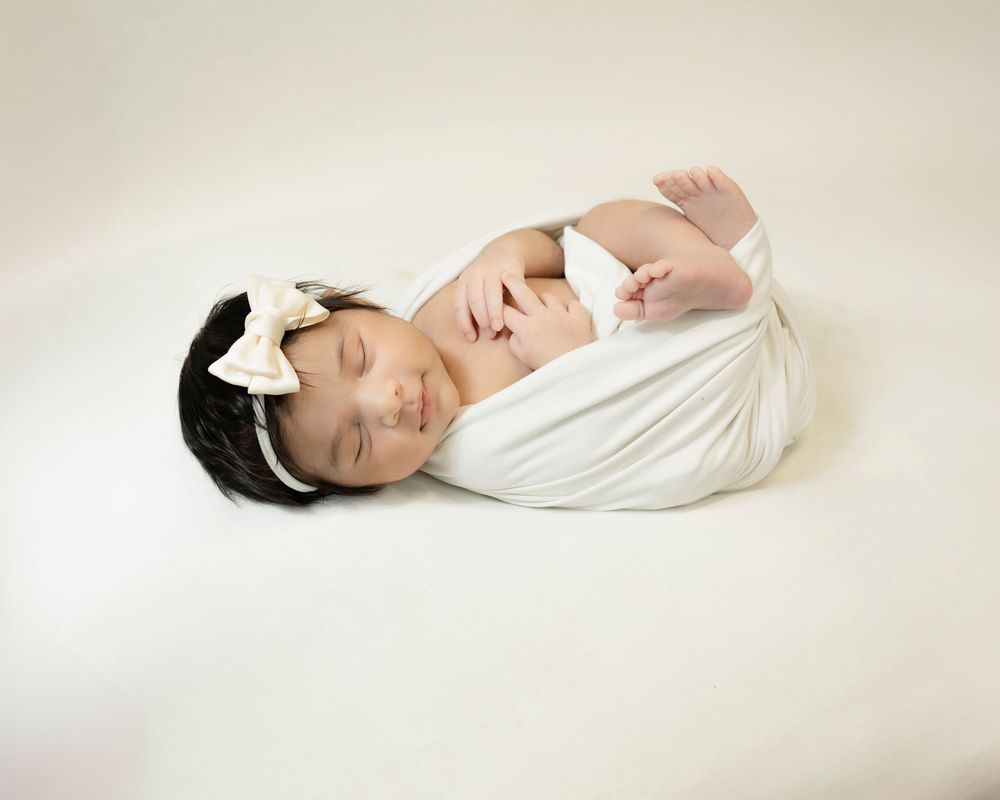 Newborn baby wrapped in soft white fabric sleeping peacefully on light background with dark headband.