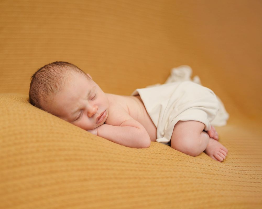 Sleeping newborn in white onesie rests peacefully on soft golden textured surface.