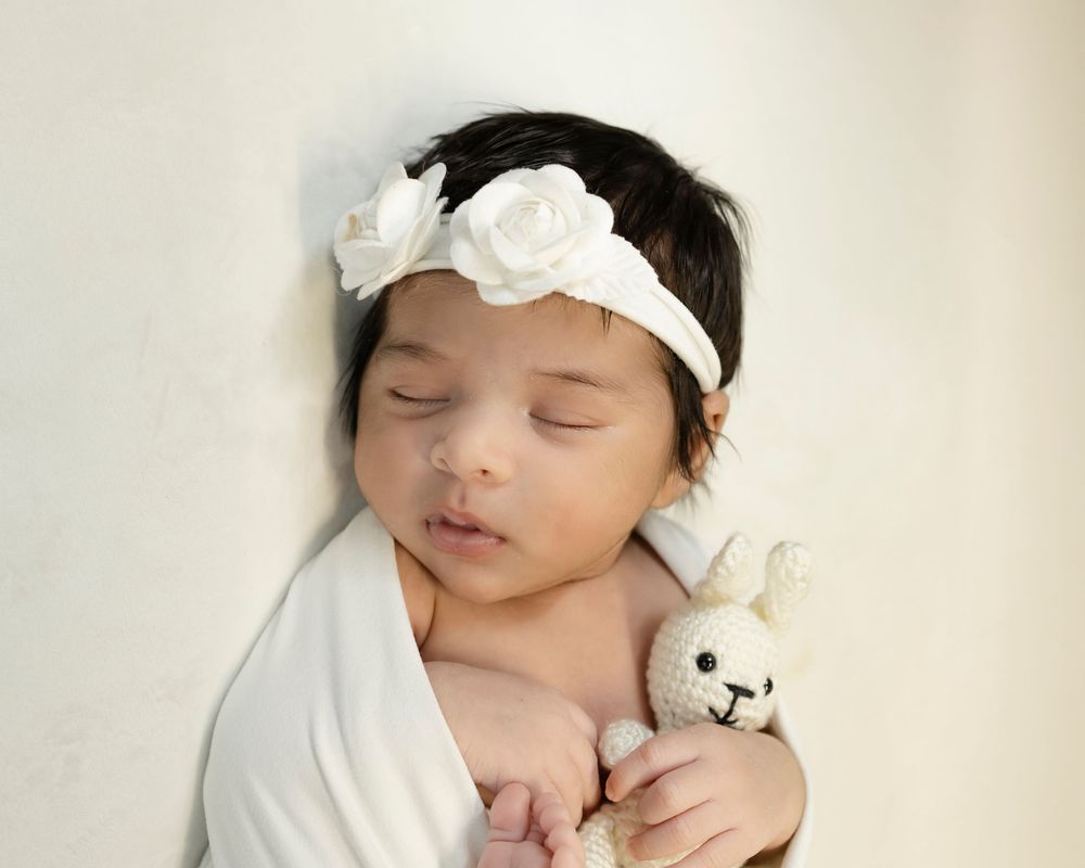 Sweet baby sleeping peacefully wearing white headband with roses while cuddling small plush toy.