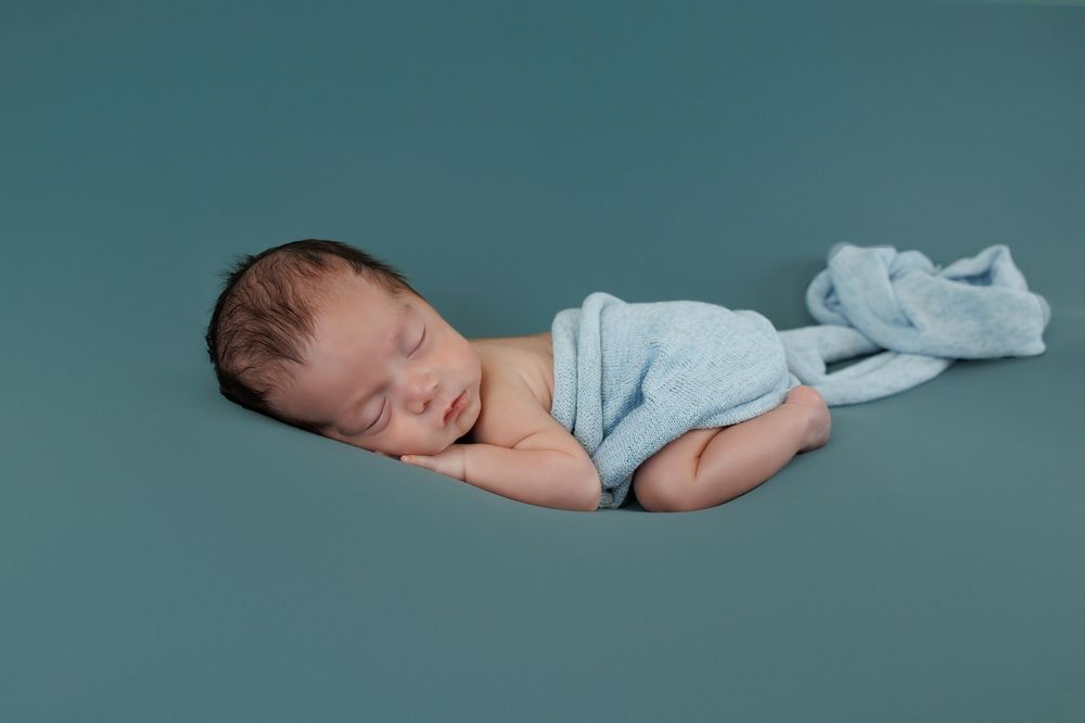 Peaceful sleeping newborn baby wrapped in soft blue blanket on teal backdrop during professional photo session.