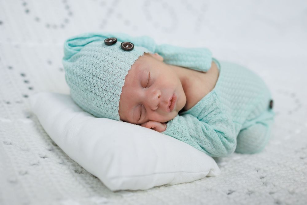 Sleeping newborn in mint green knit outfit rests peacefully on white pillow.