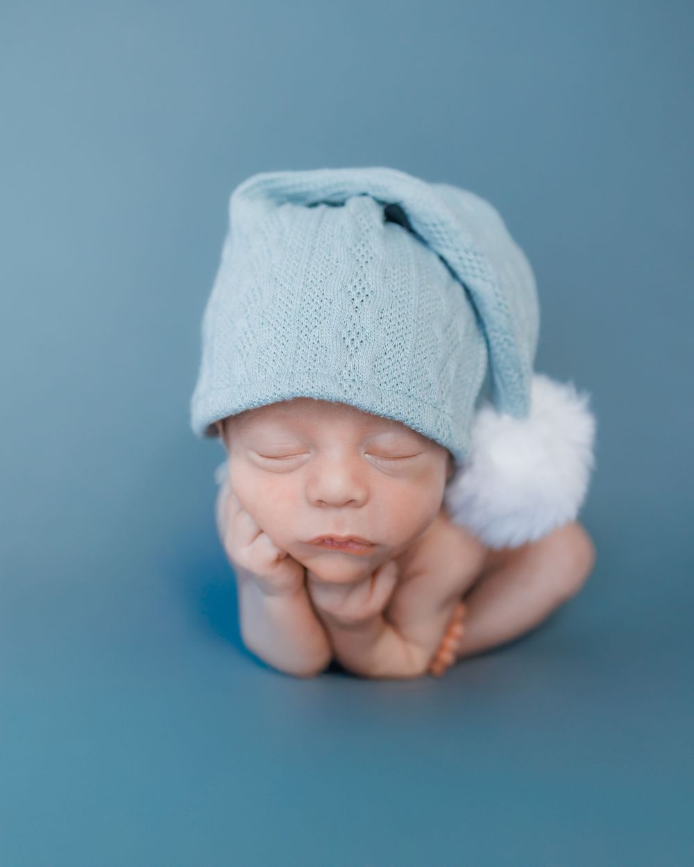 Sleeping newborn wearing soft blue knit hat with white pom pom rests chin on hands against blue backdrop.