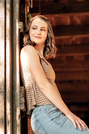 A portrait against a rustic wooden wall shows someone in a checkered top and jeans with wavy brown hair in warm lighting.