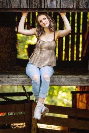 A person in ripped jeans and gingham top poses casually on a wooden deck railing in dappled sunlight.