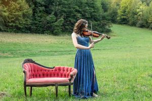 A musician in a blue dress plays violin in a grassy field with a vintage red chaise lounge.