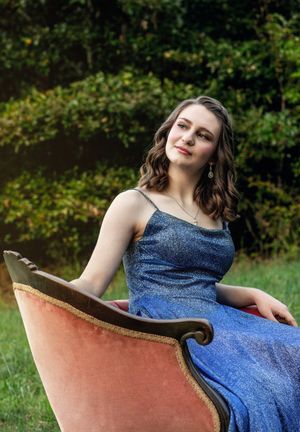 A person in a sparkly blue evening gown poses elegantly on a vintage pink velvet chair outdoors.