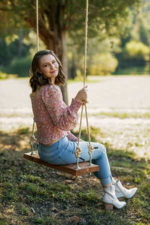 Person in floral top and jeans sits on wooden rope swing in sunny autumn garden setting.