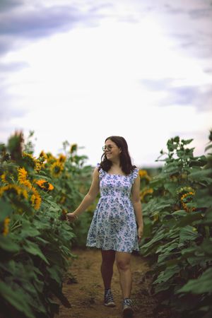 A person in a floral dress walks through a sunflower field on a cloudy summer day.