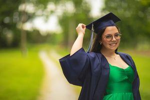 Graduate in emerald dress adjusting cap while wearing navy graduation gown outdoors.